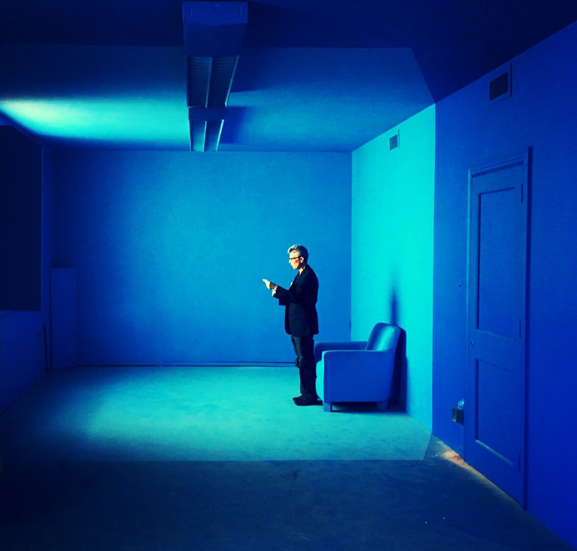 A lone figure beside a chair in a blue-lit room with a closed door.